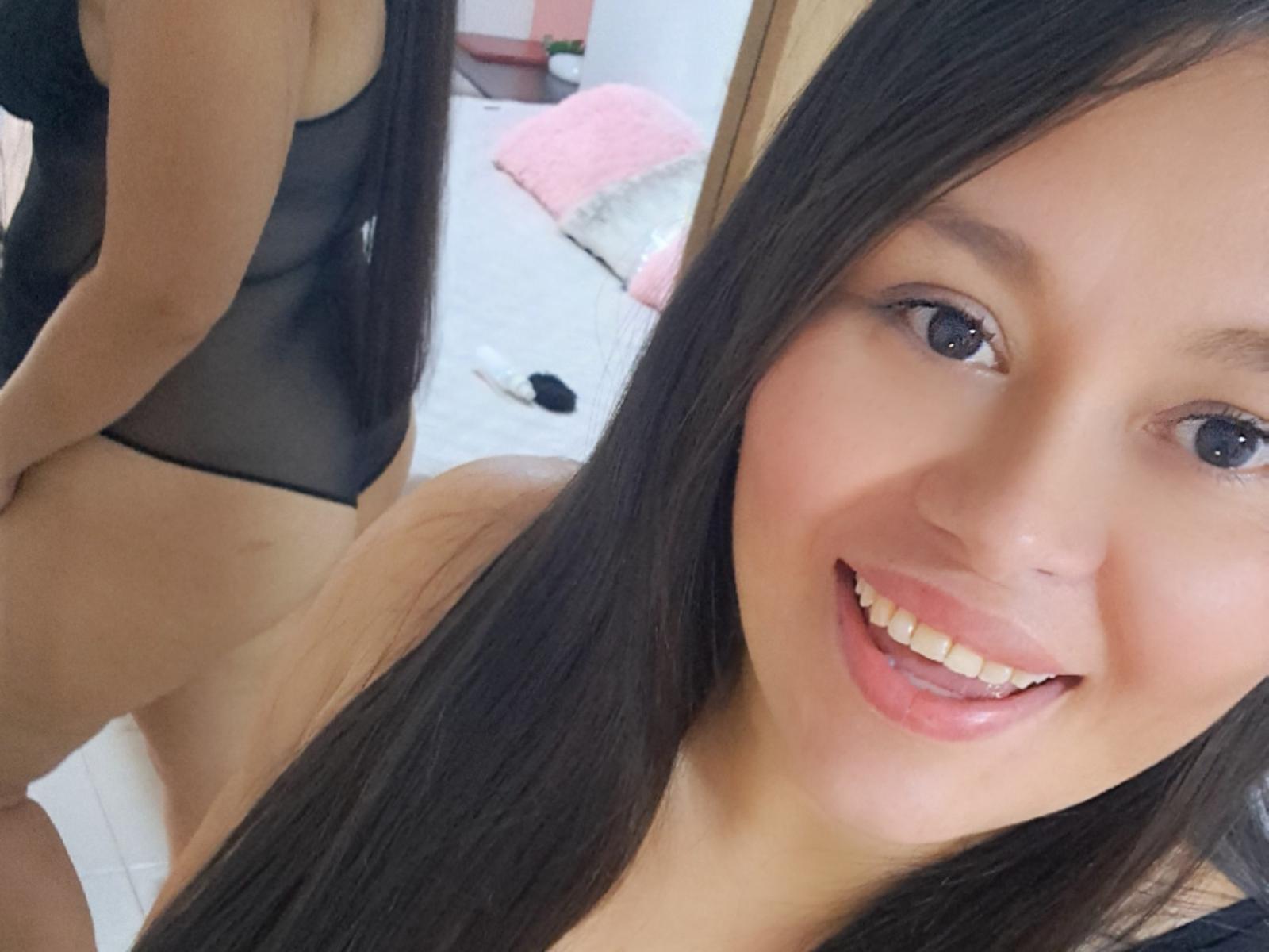 Linarodriguez with long black hair, smiling, wearing a sheer black bodysuit, showing her ass in a mirror, pink pillow, white bed, and wooden door in the background.