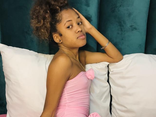 ClaraMagik with curly brown hair, tan skin, and small tits, wearing a pink strapless dress, sitting on white pillows, hand on head, looking slightly annoyed, against dark green velvet.