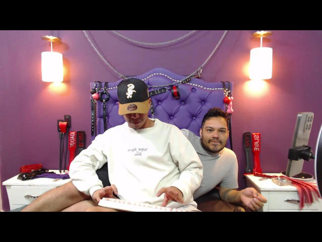 StivensAndWilliams with a black cap, white sweater, and exposed white underwear, sits on a purple bed, with another StivensAndWilliams in a gray turtleneck, looking at him.