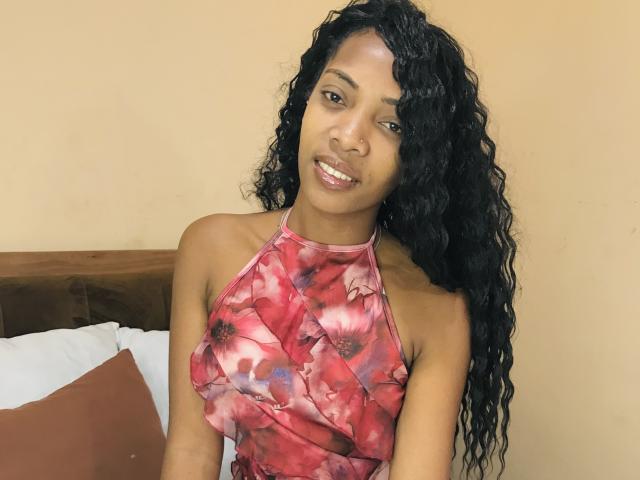 AntonniaMagik with curly black hair, wearing a red floral halter dress, smiling at the camera, showing her shiny pinkish lips and medium brown skin. Her tits are small.