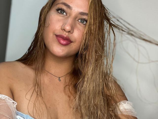 Asshly20 with wet blonde hair, pink lips, and off-shoulder white top, showing her tits. She has a necklace and a slight smile, with green eyes.