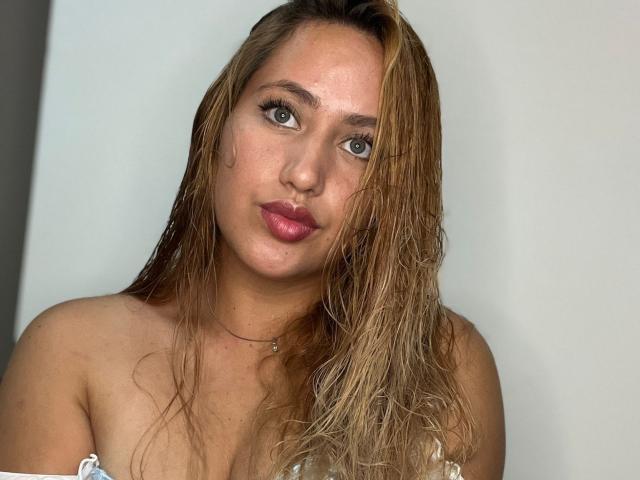 Asshly20 with wet blonde hair, full pink lips, green eyes, and a necklace, showing bare shoulders and a cute face. Boobs partially visible. Neutral expression.