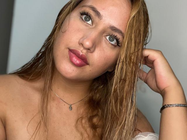 Asshly20 has wet, blonde hair, green eyes, pink lips, and a slight smile, wearing a white off-shoulder top, with a silver necklace and bracelet.