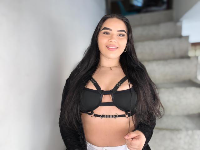 IsabellaaSweett, con lunghi capelli neri, indossa un reggiseno nero a spalline che mette in mostra il seno, e un cardigan nero, sorridendo alla fotocamera, mentre è in piedi su una scala..
