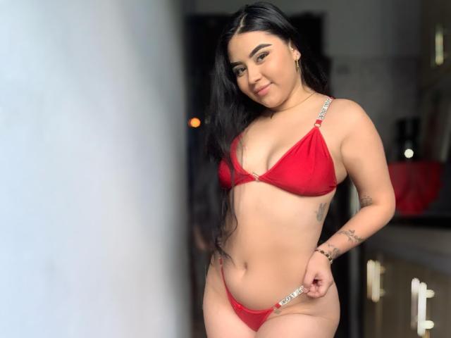 IsabellaaSweett with long black hair, red bikini, small tits, flat stomach, holding string of thong, tattoos on right arm, smiling, standing indoors, light skin, confident.