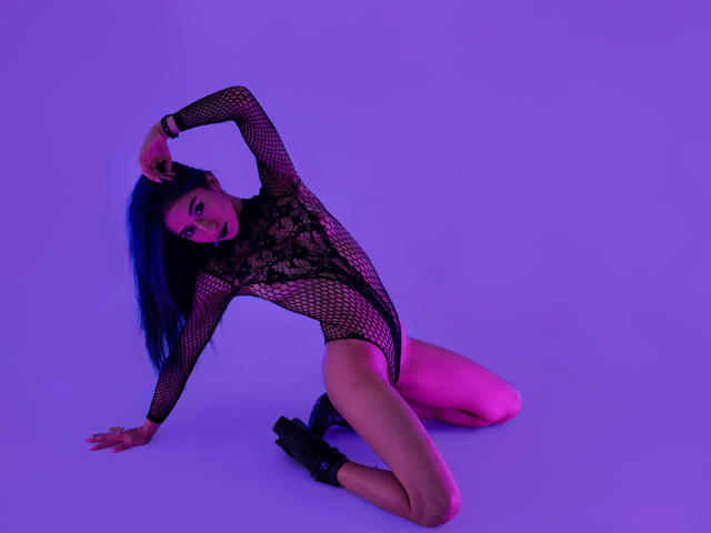 JuliaJobs is on her knees wearing a black fishnet leotard, with her tits visible through the lace. She has blue hair, a purple glow, and black boots.
