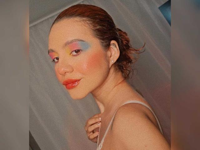 AmbeerLux with red lipstick, pink and blue eye makeup, and a slight smile, has her brown hair in a messy bun, wearing a white strap dress, and a small earring.