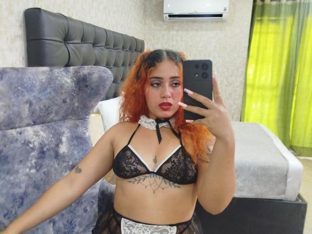 MerydaBraveI with orange curly hair, black lace bra, white choker, tattoos under boobs, holding phone, mirror selfie, red lipstick, bed with gray headboard, green curtain.