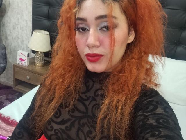 MerydaBraveI, con cabello naranja rizado, una blusa de encaje negro, lápiz labial rojo y maquillaje de ojos sutil, sentada en una cama, sonriendo ligeramente, mostrando sus pechos..