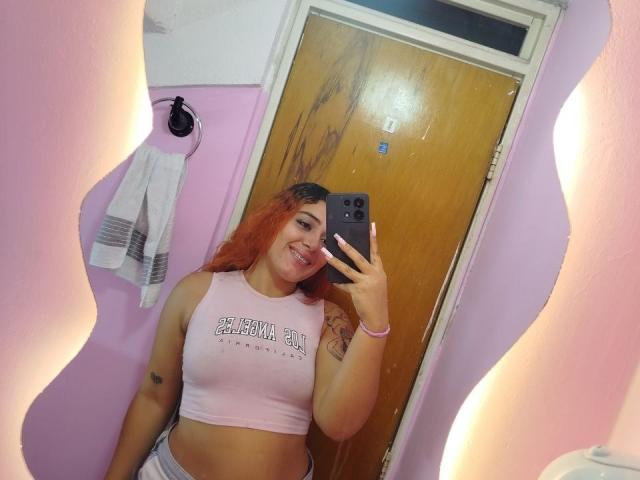MerydaBraveI with orange hair, smiling, holding phone, wearing pink crop top, showing tits, tattoo on arm, mirror selfie, pink walls, wooden door, towel on wall.