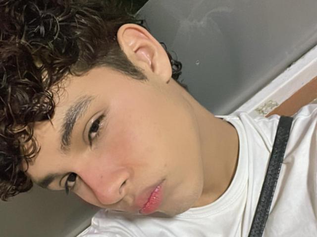 DenisBachh with curly dark hair, light skin, and pouty pink lips, wearing a white shirt with a black strap, looking down with a slight frown.