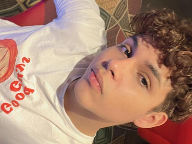 DenisBachh with curly brown hair, light skin, and full lips, wearing a white shirt, looking up with a slight pout. Red chair in background. Slight shadow on face.