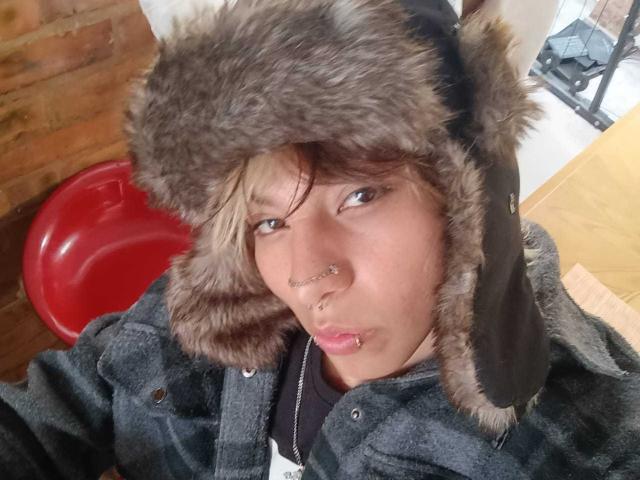 OliIvero, con un cappello marrone e peloso, una giacca nera, un anello al naso e un piercing al labbro, che sembra leggermente annoiato. Un oggetto circolare rosso sullo sfondo. Pelle marrone chiara, capelli biondi corti..
