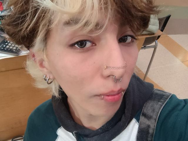 OliIvero hat kurze, lockige blonde und braune Haare, trägt mehrere Piercings (Nase, Lippe, Ohr) und ein grün-weißes Hemd, und zeigt auf einem Selfie einen ernsten Gesichtsausdruck..