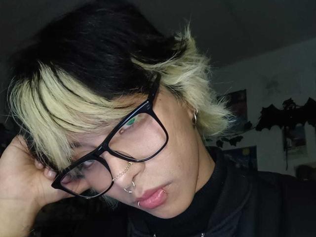 OliIvero with black-and-blonde hair, black glasses, nose ring, and lip ring, looking down with hand in hair. OliIvero's face is slightly shadowed, wearing a black top.