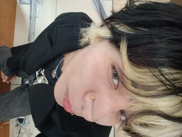 OliIvero, avec des cheveux noirs et blonds, des piercings au nez et une veste noire, regardant vers le haut avec des yeux bleus, portant un collier en chaîne, dans une salle de bain..