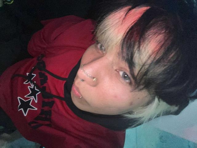 OliIvero, com cabelo rosa e preto, piercings no nariz e no lábio, vestindo uma camisa vermelha com estrelas pretas, olhando para a câmera..