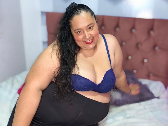 TistMature tiene senos grandes, lleva un sostén de encaje azul, pantalones negros y tiene una sonrisa. Tiene curvas, cabello negro largo, lápiz labial rojo y, en el fondo, un cabecero rosa con borlas..