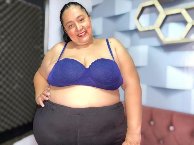 TistMature is a chubby woman with dark hair, wearing a blue lace bra, showing her big boobs, and black pants, smiling with red lipstick in the photo.