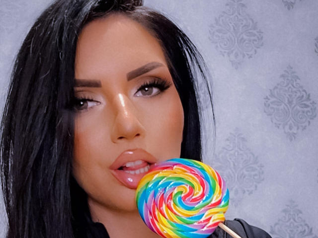 PaolaAdams with black hair, brown eyes, and glossy pink lips, sucking on a colorful swirl lollipop, her tongue partially out, showing off her shiny, wet-looking lips.