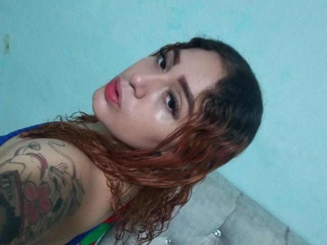 MerydaBraveI with wet brown hair, pink lips, and tattoos on her shoulder, looking over her shoulder at the camera. Her boobs are partially visible in a colorful top.