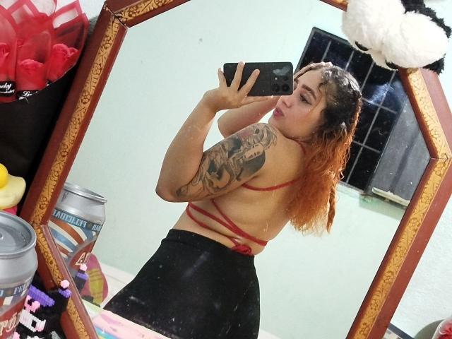 MerydaBraveI with orange hair, tattoos on left arm, red strappy top, black skirt, taking selfie in mirror, showing back and butt, holding phone.