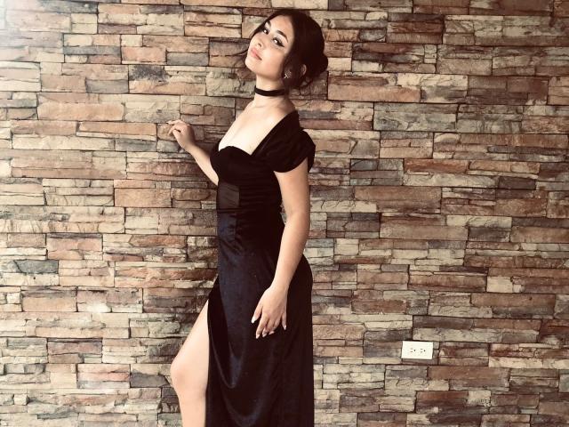 BellaMiaI with big tits, wearing a black dress with a thigh-high slit, choker, and updo, standing against a stone wall, looking back with a slight smile.