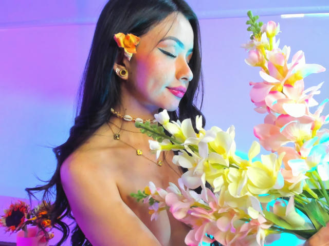 KattRouse with long black hair, orange flower in hair, gold earrings, necklaces, holding pink and white orchids, pink and blue lighting, smiling, topless, with small tits.