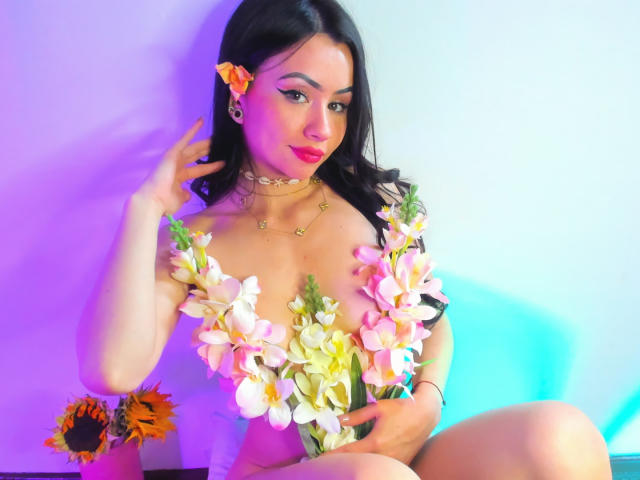 KattRouse with dark hair, pink flowers covering her tits, wearing gold necklaces, sitting with a hand by her face, orange flower in her hair, and sunflowers on the left.