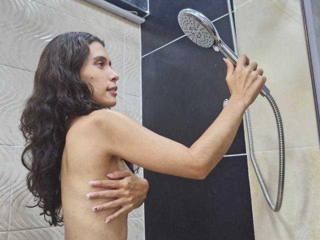 KatheKiss with long curly black hair, holding a showerhead, covering her small bare tits with one hand, water spraying, looking to the side in a modern shower.