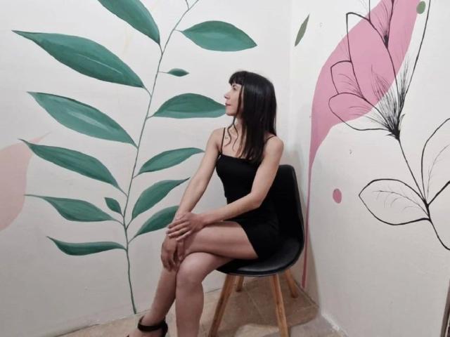 Milagros, una mujer atractiva con cabello negro, senos pequeños y un mini vestido negro, está sentada en una silla, con las piernas cruzadas, en una habitación con decoración de hojas verdes y flores rosas en las paredes..