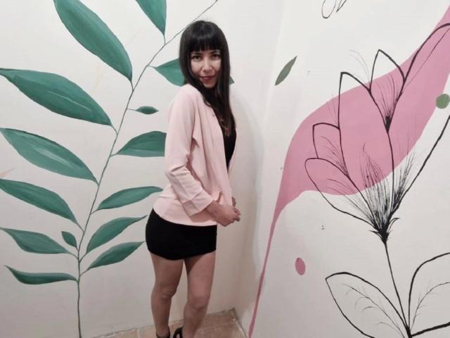 MilagrosPop, attraente con i capelli neri, un cardigan rosa e pantaloncini neri, in piedi con le mani sui fianchi, sorridente, che mostra il sedere, in una stanza con decorazioni murali a tema foglie verdi e fiori rosa..
