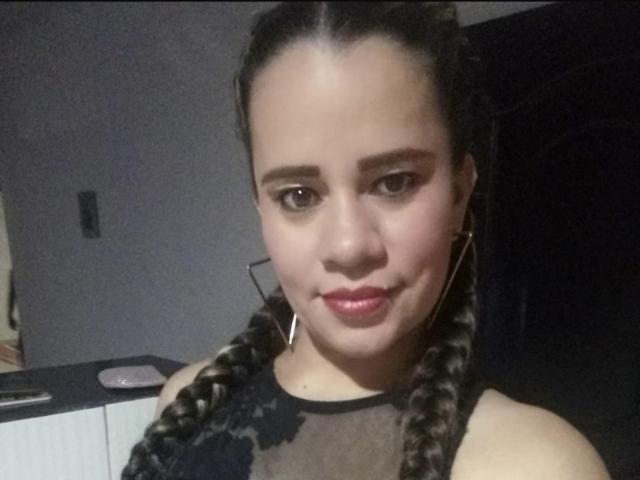 CharllotI with braided hair, medium skin, brown eyes, and red lipstick, wearing a black sheer top, showing a slight smile, and wearing gold earrings.