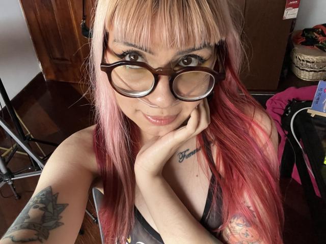 KallySpice with pink and blonde hair, glasses, and tattoos, smiling and touching her cheek, showing her tits in a black tank top.