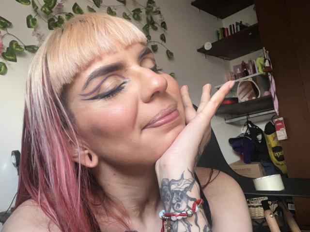 KallySpice with blonde and pink hair, eyeliner, and tattooed hand, smirking with eyes closed, resting chin on hand, showing off pierced ear and bracelet.