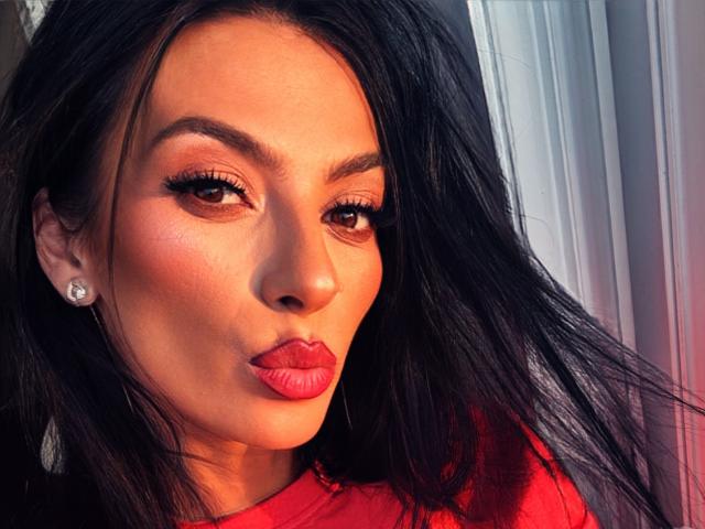 KinkyAmelie's face with dark hair, pink lipstick, and earrings, pouting her full, shiny lips. She's wearing a red top, with warm sunlight highlighting her glowing skin.