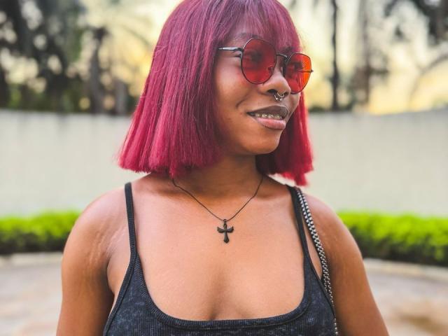 amberryider with red bob, nose ring, red sunglasses, black tank top, cross necklace, smiling, showing teeth, black lipstick, holding black and white bag, outdoor setting.