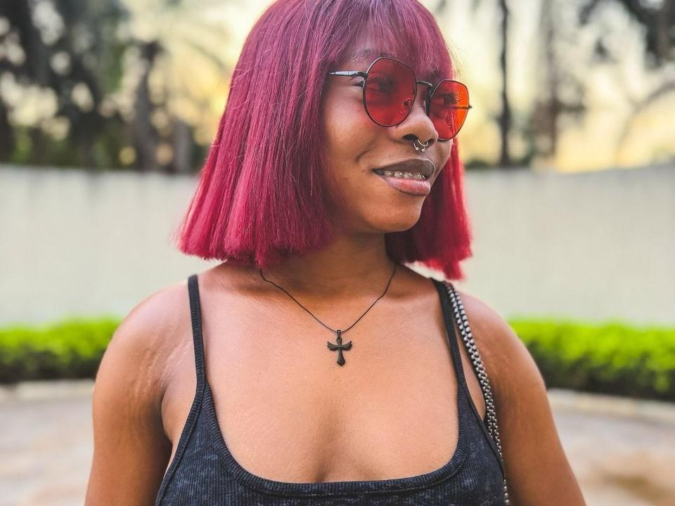 amberryider with red bob, nose ring, red sunglasses, black tank top, cross necklace, smiling, showing teeth, black lipstick, holding black and white bag, outdoor setting.