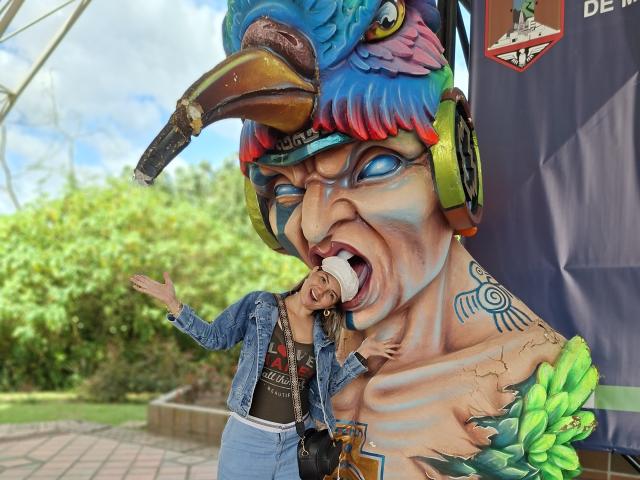 KatherineToro with her tongue in a giant painted face with a bird mask, colorful feathers, and blue eyes, wearing a denim jacket and jeans, in front of a large mural.