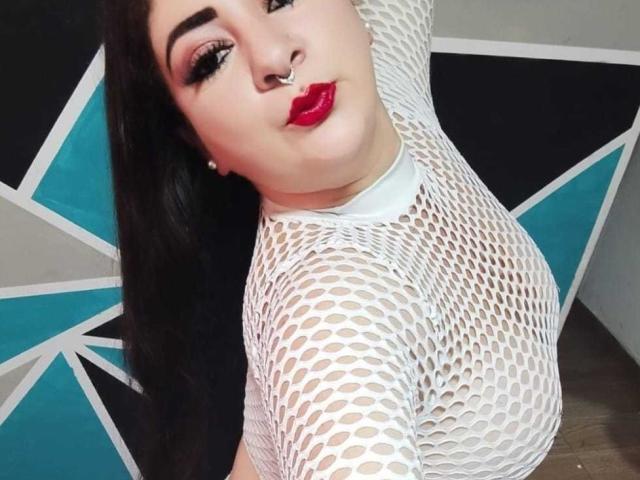 ZoeMonroe with dark hair, red lipstick, and nose ring, wearing a white fishnet top, showing her tits through the mesh. She's looking up, with a slightly pouty expression.