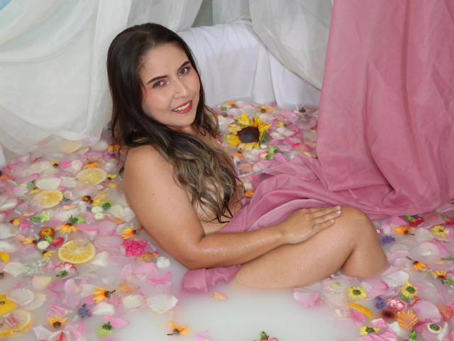 SakuraBlaze with wet skin, long brown hair, and pink cloth covering her ass, sitting in a flower-filled bathtub, smiling at the camera. Sunflower in water, pink curtain.