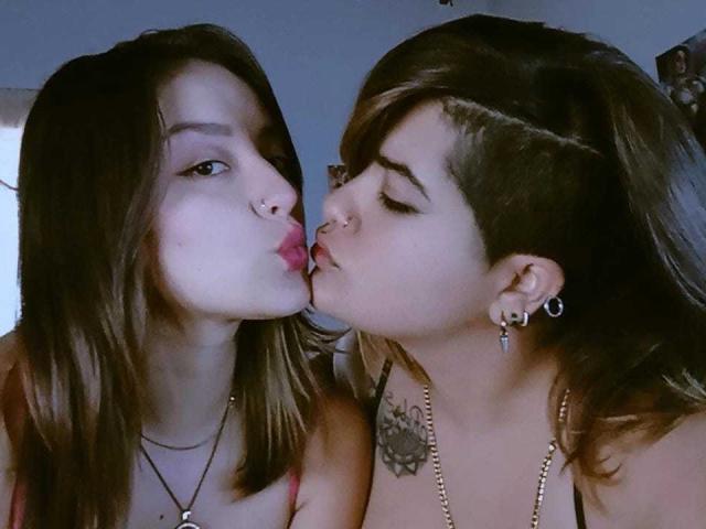 ViolletAndSussan are kissing with pink lips, Viollet has nose ring and long brown hair, Sussan has undercut, earrings, tattoo, and brown hair, both wearing necklaces.
