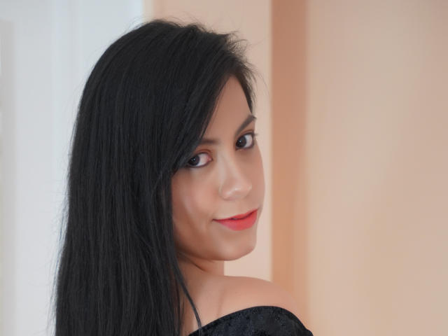 LunaFontana with black hair, red lipstick, and a slight smile, looking over her shoulder, showing off her bare shoulder and a hint of her black top.