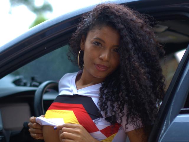 AnelicaSexos with curly dark hair, wearing a white and red geometric shirt, holding a box, leaning on a car door, smiling, large hoop earrings, medium brown skin.