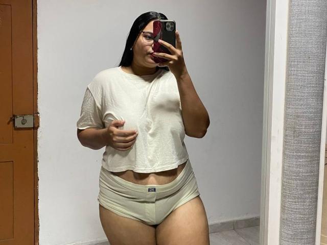 NinaBigTitsHot with wet shirt, cream-colored panties, and dark hair, taking a mirror selfie, showing her tits and ass, holding phone with red case, in a room.