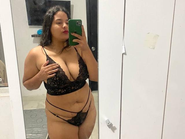 NinaBigTitsHot with dark hair, red lipstick, black lace bra and panties, holding green phone, mirror selfie, curvy, medium skin, visible cleavage, hand on chest, black straps on panties.