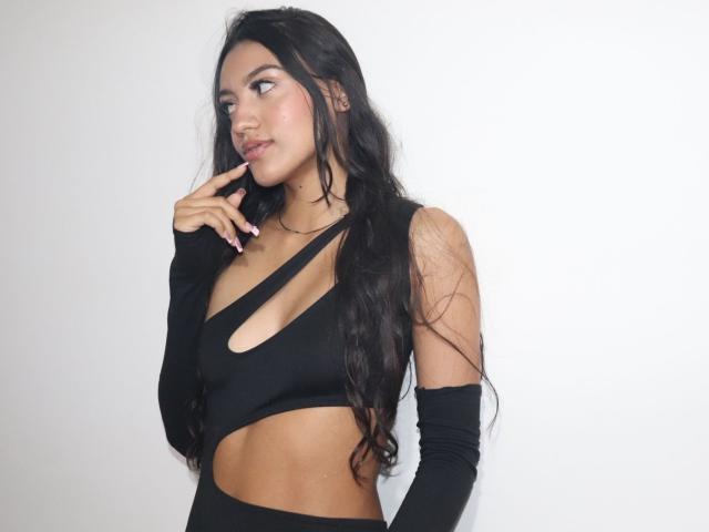 MiaaRyder with long black hair, wearing a black cutout top and arm sleeves, biting her finger, showing off her small tits and midriff, looking to the side.