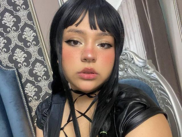 AshMorgana, avec ses cheveux noirs, sa frange et son eye-liner, porte un haut en cuir noir avec un décolleté à lacets, a un anneau de nez et des lèvres roses, et est assise sur une chaise en argent..
