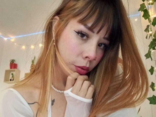 AriiannaHot with pink nose, black eyeliner, and pink lipstick, touching her chin with a hand, wearing a white top, pearl necklace, and blonde hair with brown roots.