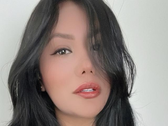 AlessandraMalory has black, wavy hair covering one eye, with light brown skin, subtle makeup, and glossy pink lips. She's looking slightly to the side with a small smile.